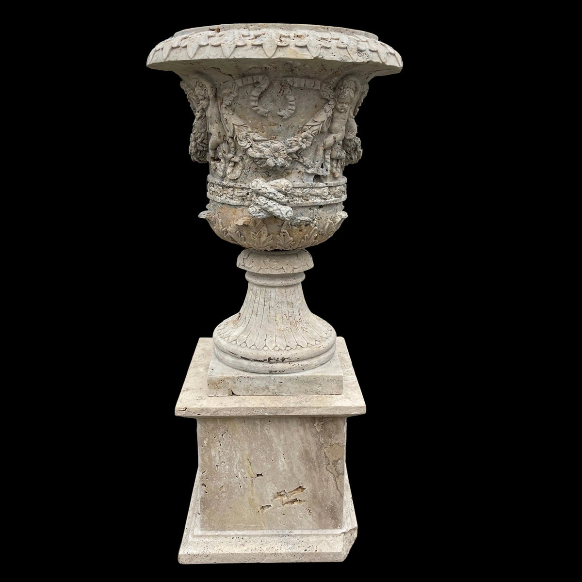 A large carved stone garden urn with a matching pedestal base, photographed outdoors on a concrete surface with a fence, trees, and a small shed in the background.