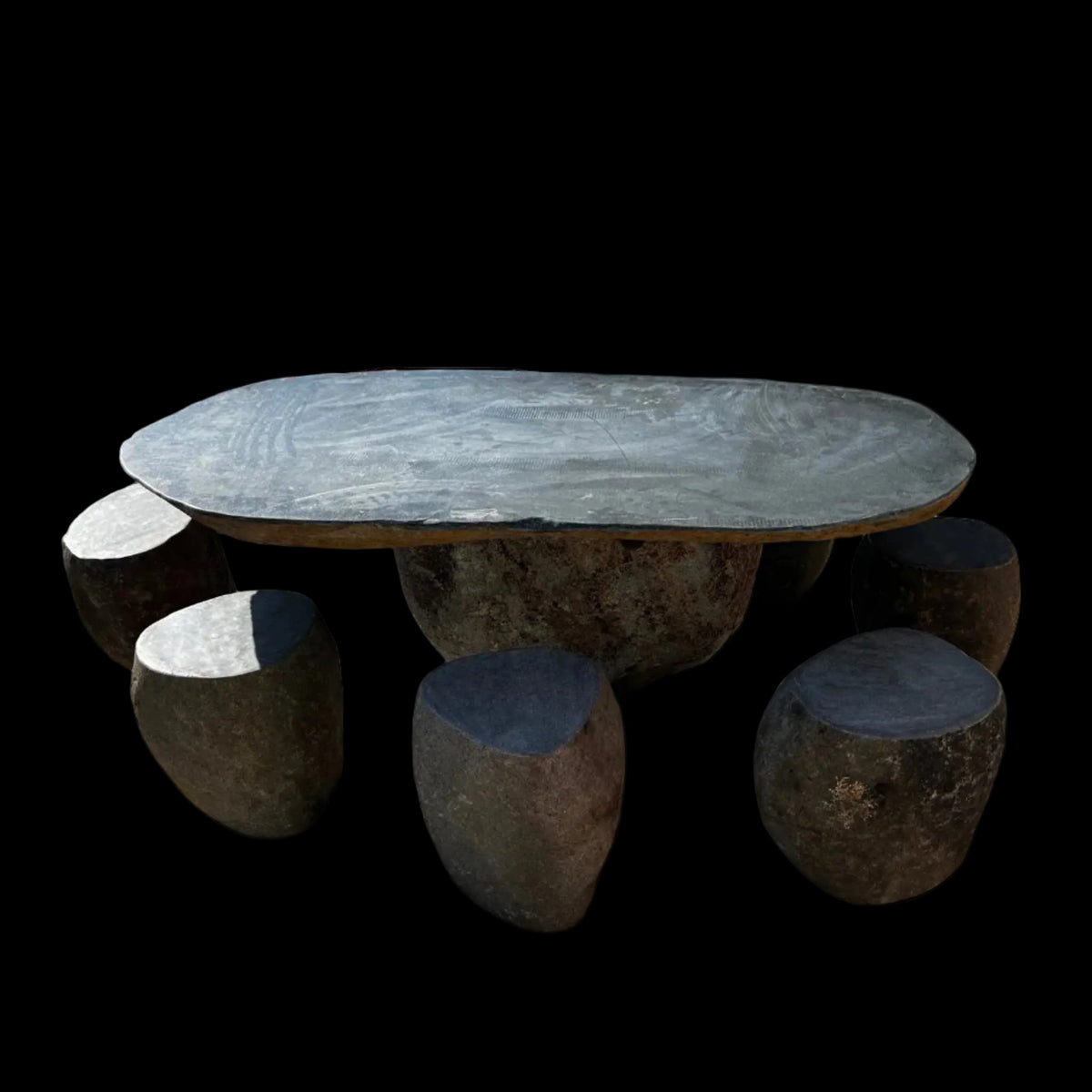 A natural granite stone 6-seater table with a round top and eight uniquely shaped granite stools placed on a tiled surface with a brick wall in the background.