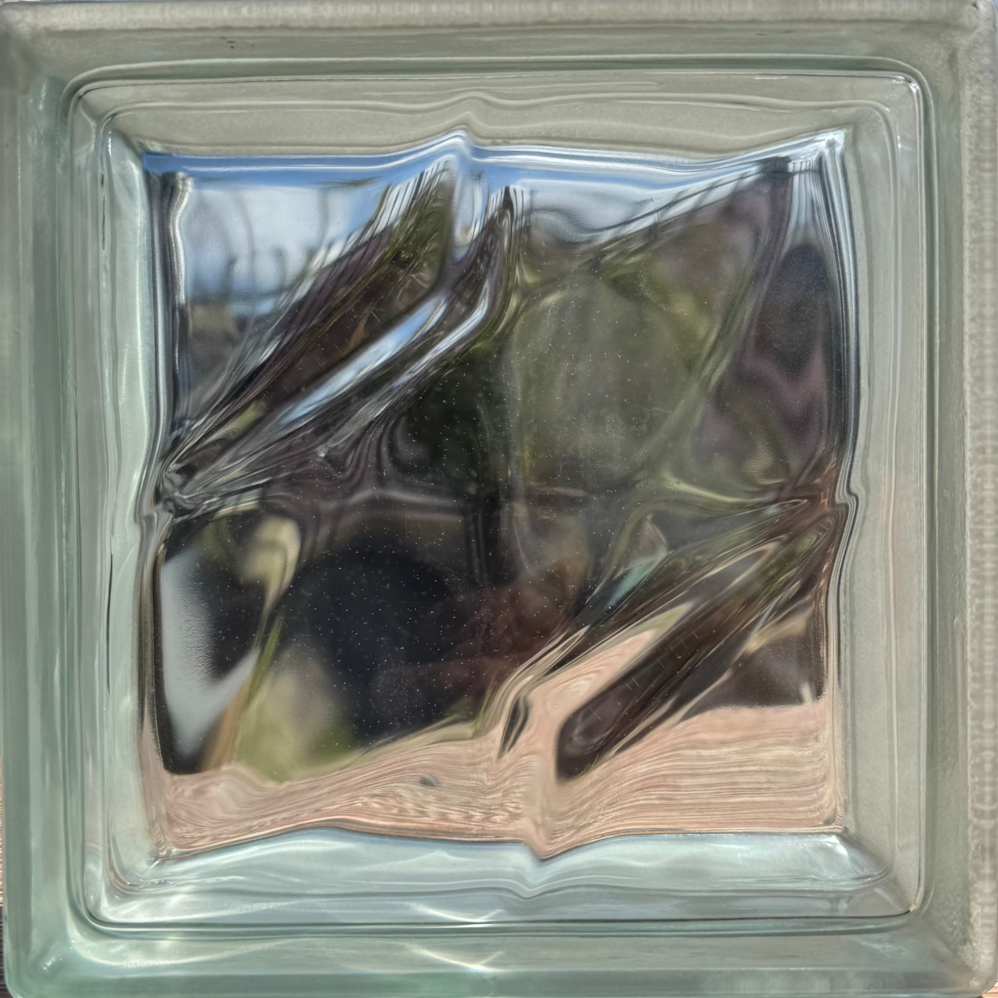 A square glass block window with a textured, wavy pattern of light and reflections inside, framed by a clear glass border.