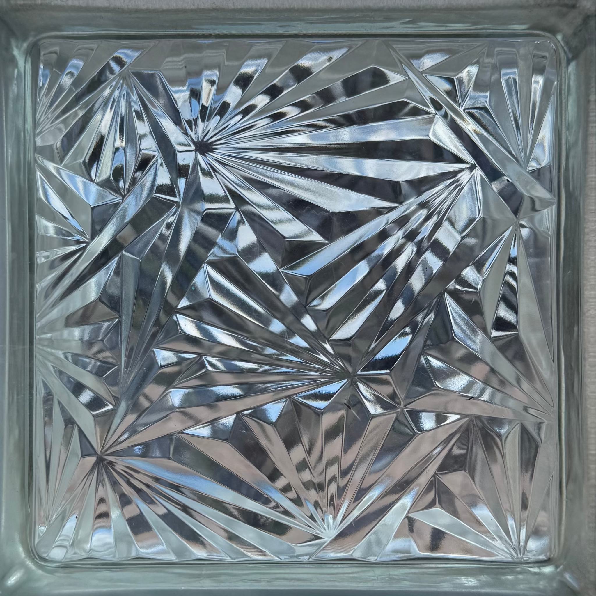 A square clear glass block with an etched starburst and geometric faceted pattern on the front, shown against a light neutral background.
