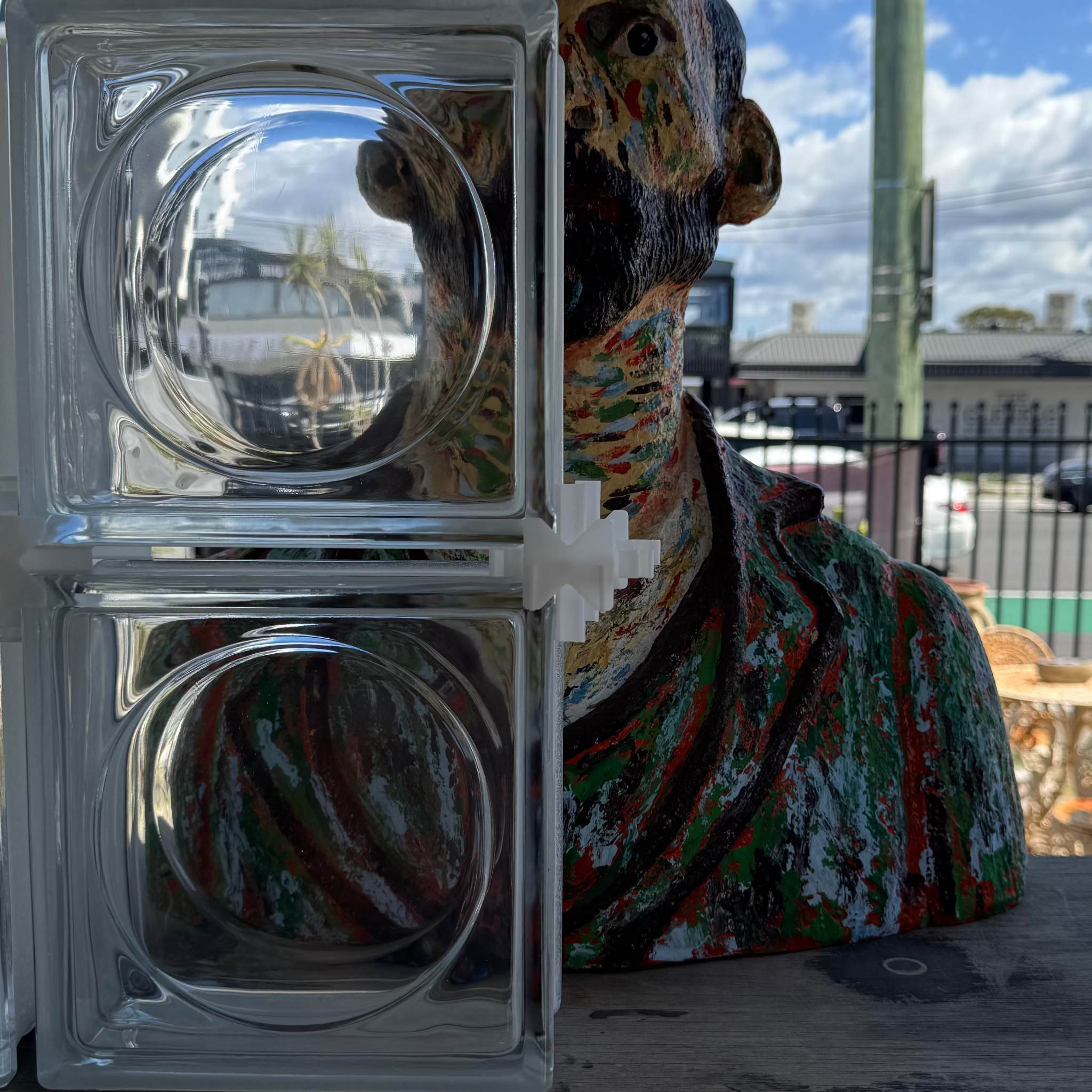 A clear glass block window or wall panel with two circular glass inserts, shown next to a colorful painted bust sculpture in an outdoor setting.