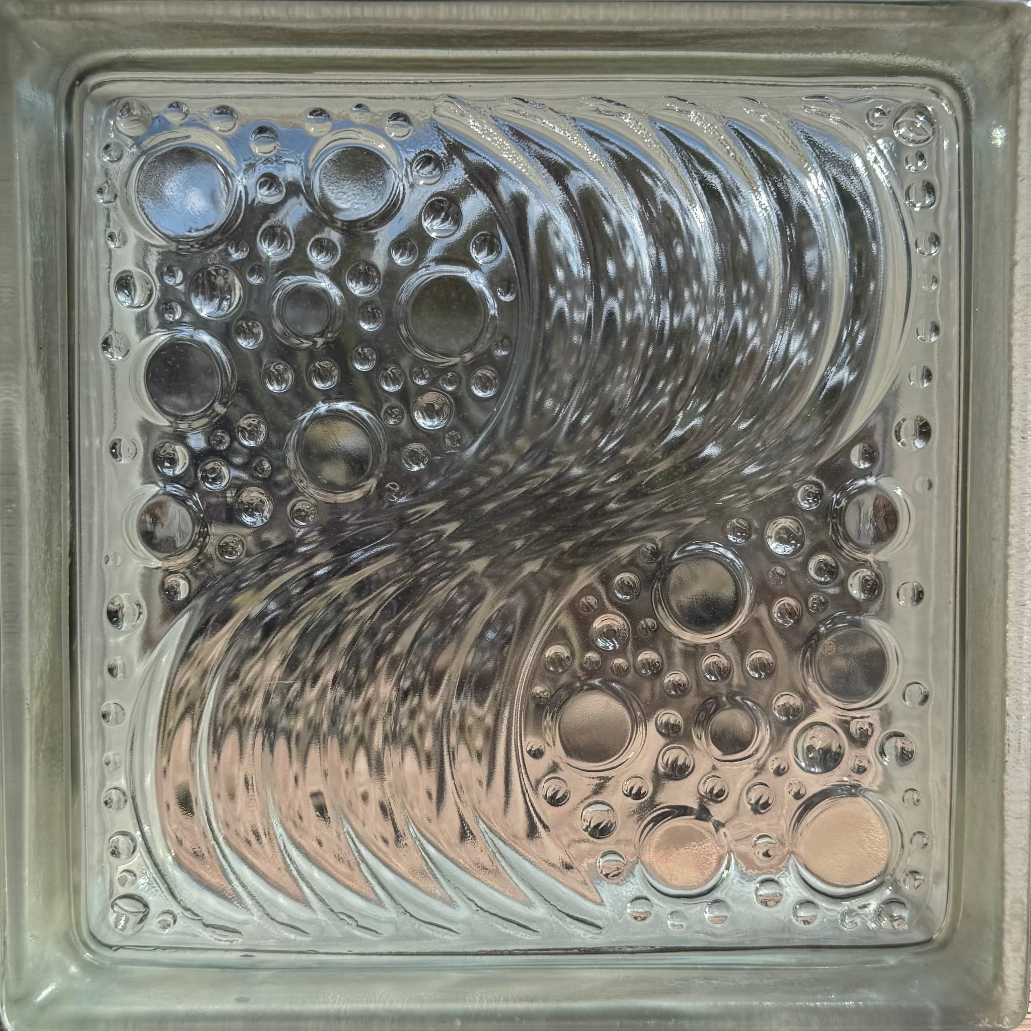 A square glass block featuring an embossed, reflective wave design with circular bubble-like patterns in a metallic silver finish.