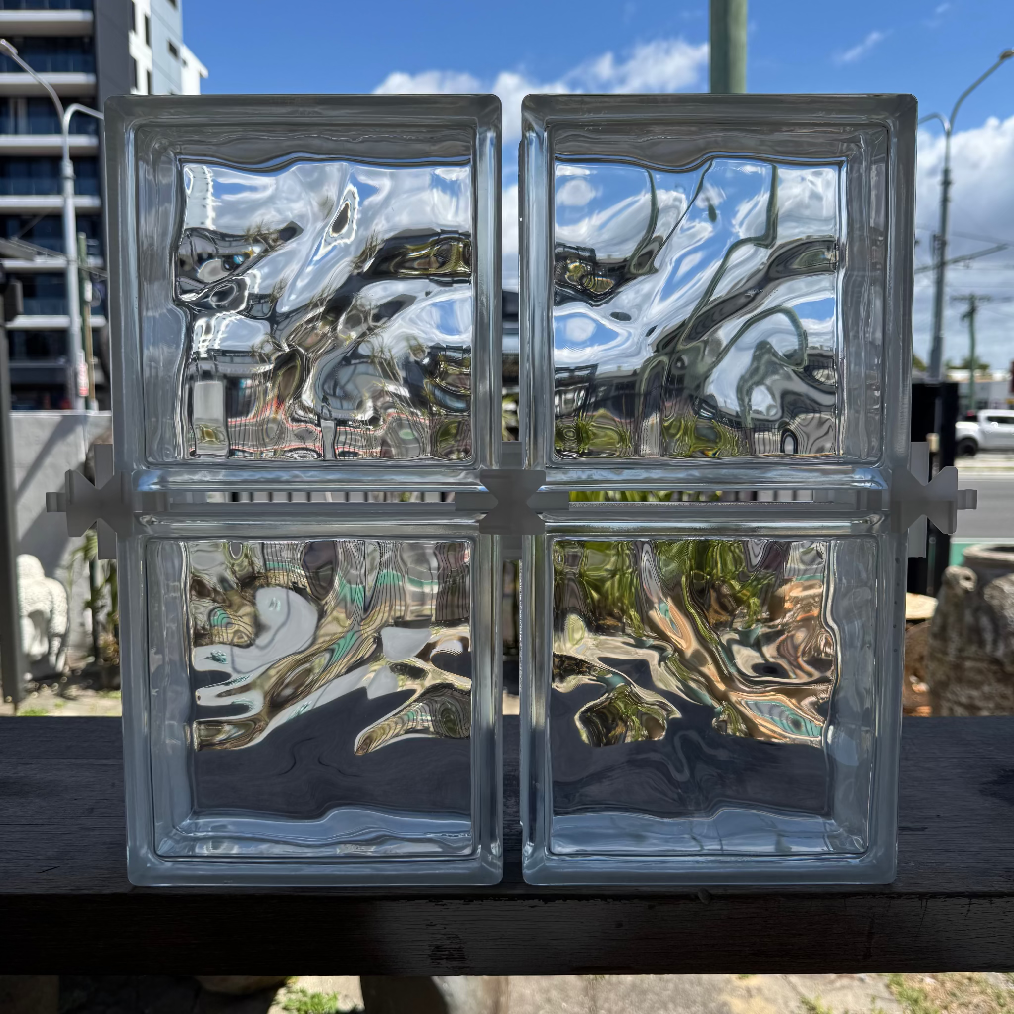 Two square glass blocks with a wavy, rippled texture, arranged side-by-side on a dark wooden surface outdoors under a blue sky.