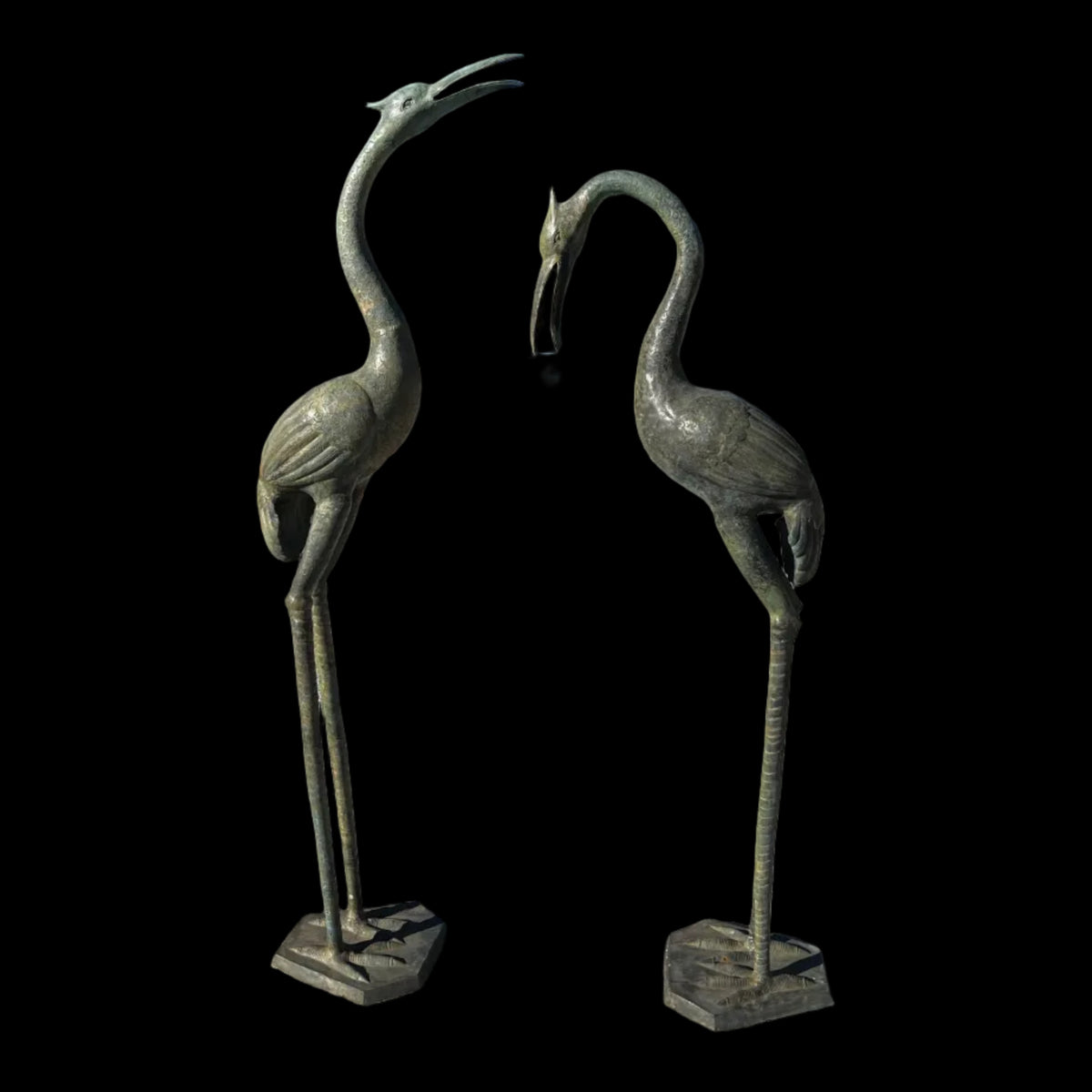 Two bronze metal stork sculptures with a copper patina finish standing on a concrete ground with a blue sky in the background.