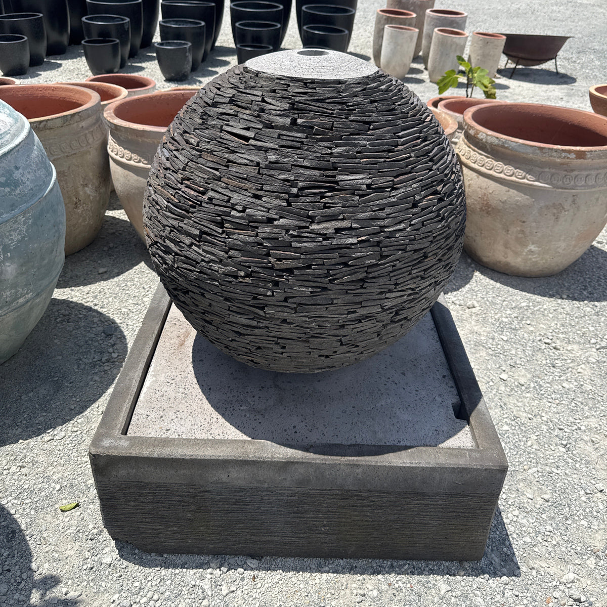A 2 piece inlaid shale rock water fountain with a sphere on top flowing into a concrete base, displayed outdoors.