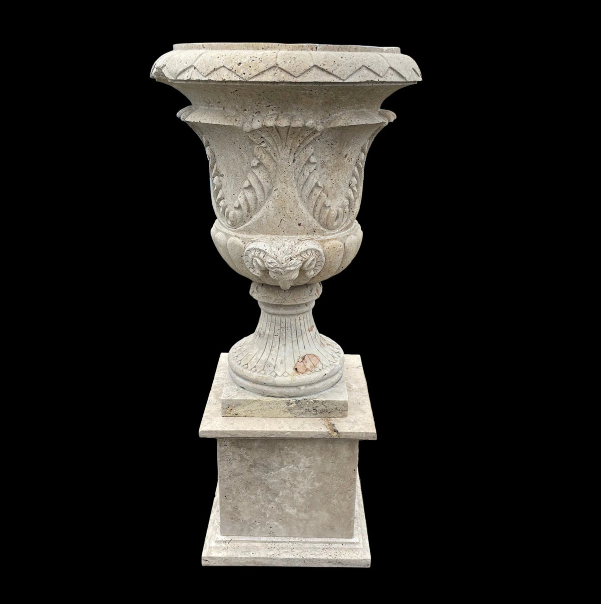 A large carved stone planter urn on a matching square pedestal base, photographed outdoors on a paved lot with a blue truck and buildings in the background.