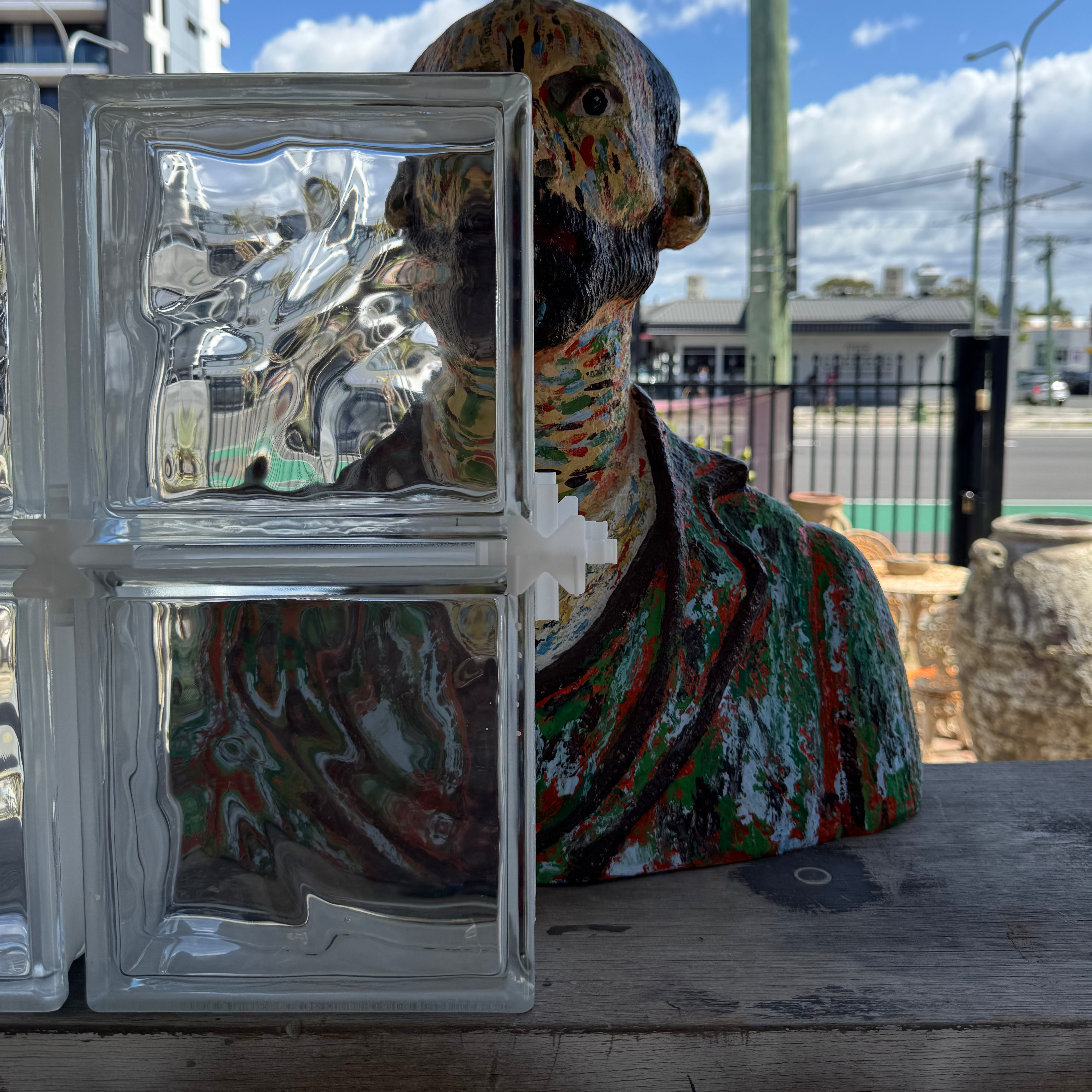 A colorful painted bust sculpture of a bald man wearing a robe, displayed outdoors on a wooden table next to clear glass blocks under a blue sky.