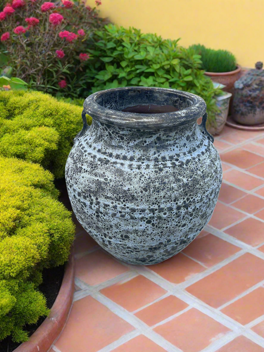 Lava rock Atlantis garden pot urn with handles in beige colour on terracotta patio next to a beautiful garden