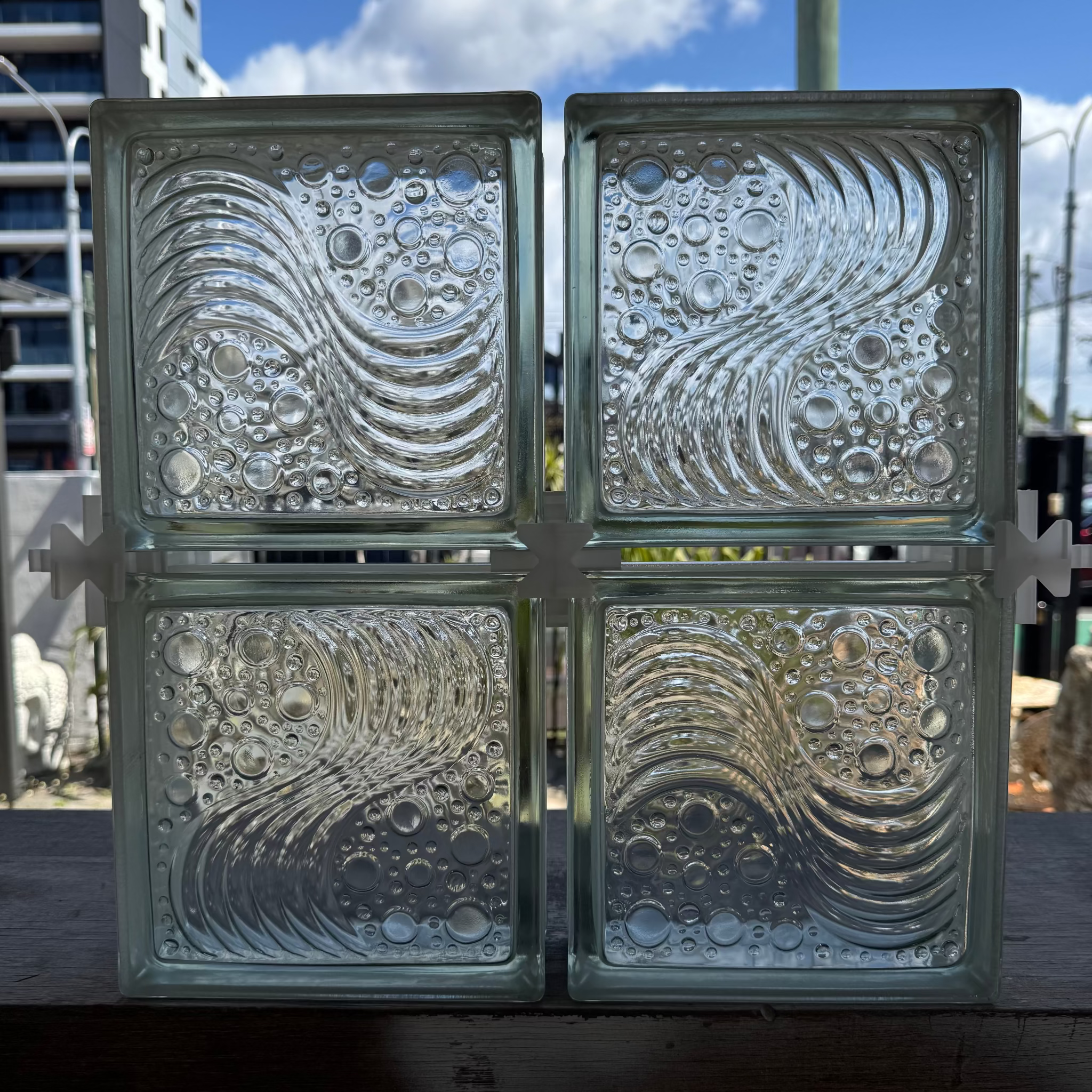 Four clear glass blocks with embossed wavy and bubble patterns, arranged in a 2x2 grid, photographed outdoors on a wooden surface with a city street and blue sky in the background.
