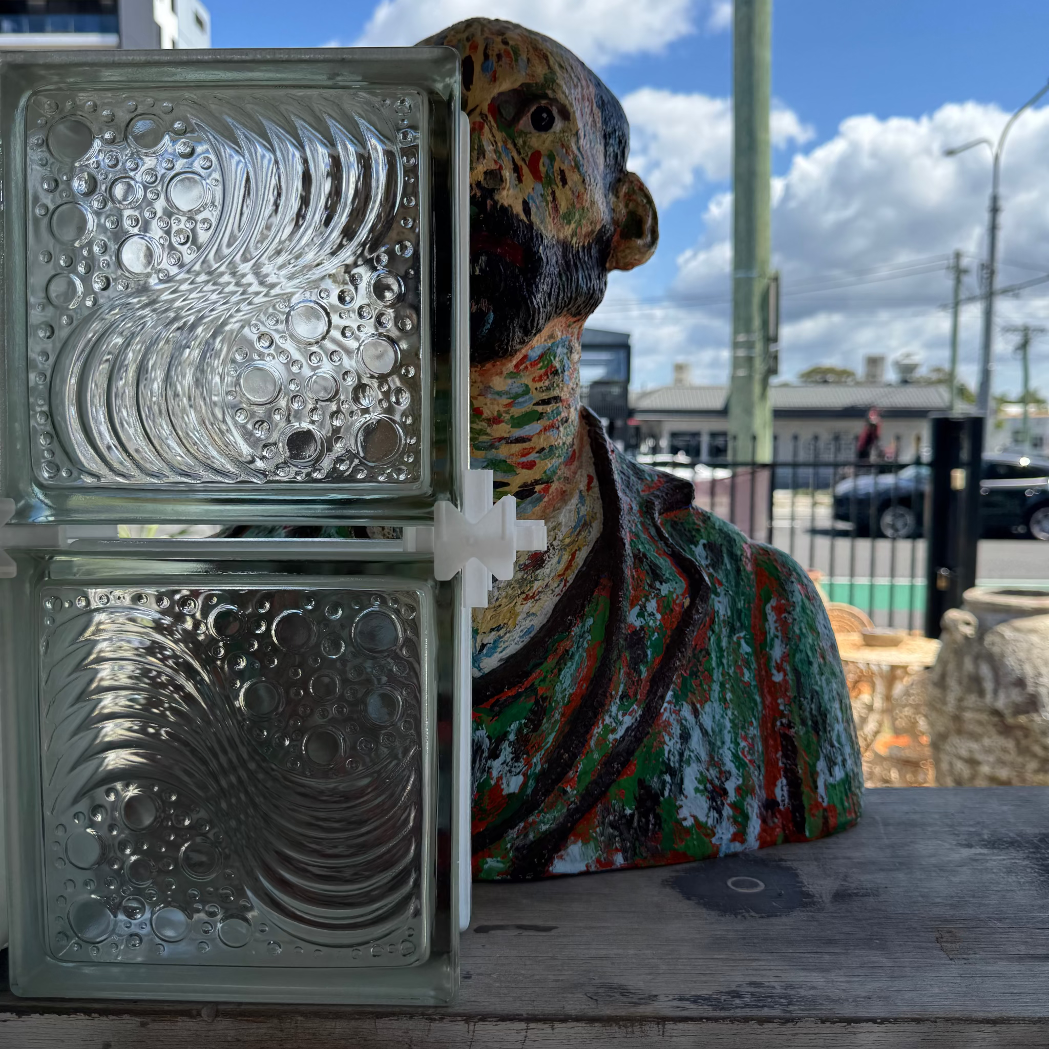 A decorative glass block wall light with a colorful painted bust sculpture positioned behind it on an outdoor wooden surface, with a street scene and blue sky in the background.
