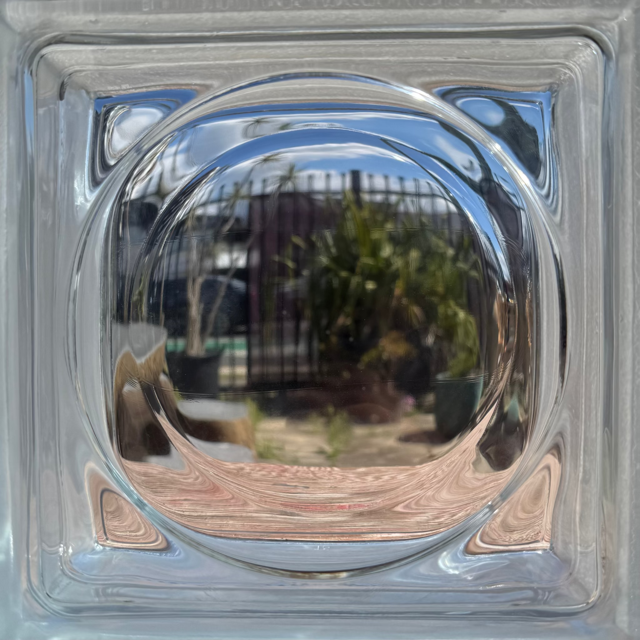 A square clear glass block with a raised circular center, photographed from the front. The transparent glass reflects an outdoor scene with plants, a fence, and sky.