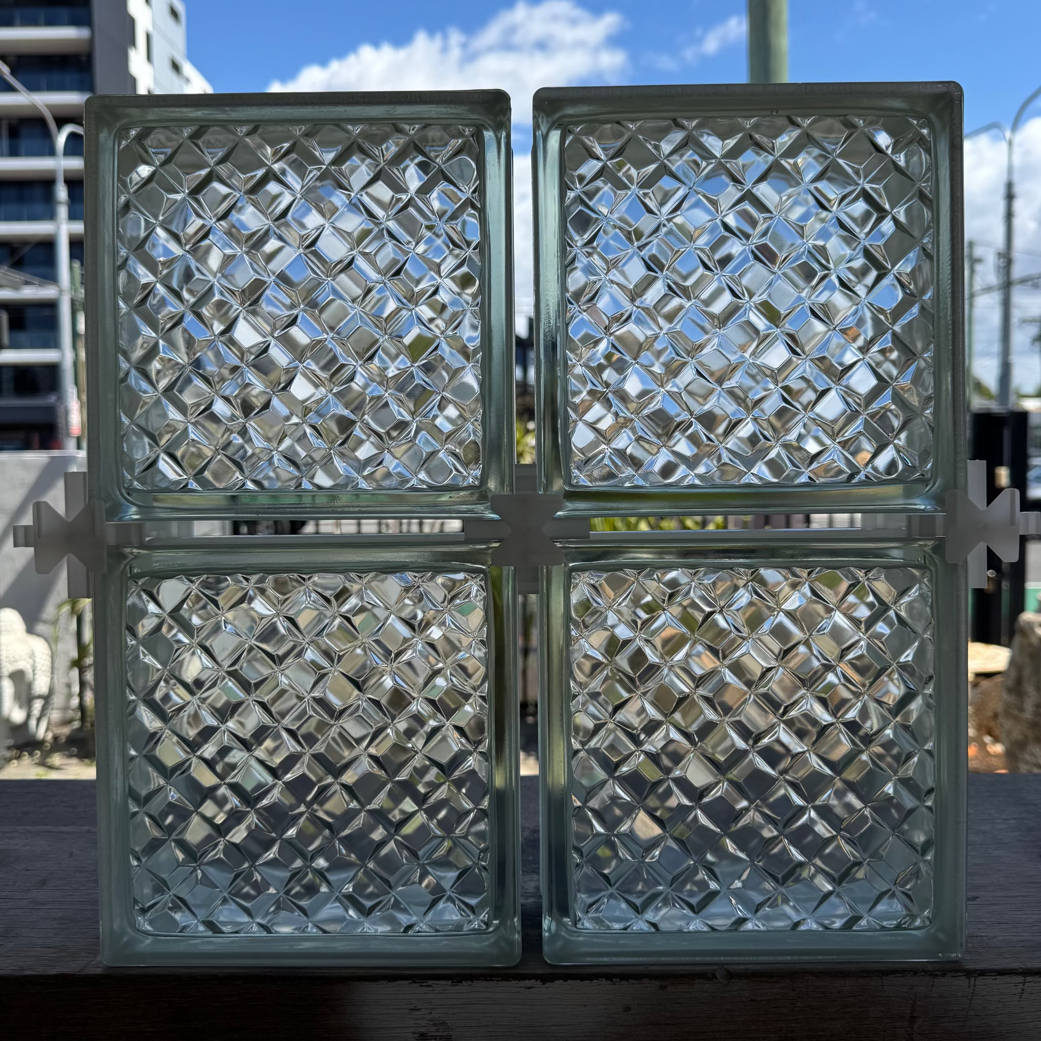 Four square glass block panels with a diamond-patterned embossed design, arranged in a 2x2 grid on a wooden surface outdoors.